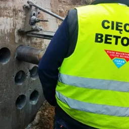 Pracownik w kamizelce odblaskowej z napisem 'Cięcie Betonu' wierci otwory w betonowej ścianie za pomocą specjalistycznego wiertła rdzeniowego.