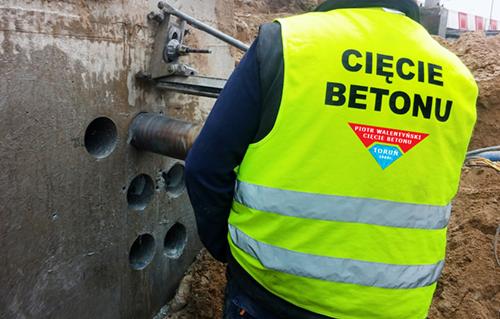 Pracownik w kamizelce odblaskowej z napisem 'Cięcie Betonu' wierci otwory w betonowej ścianie za pomocą specjalistycznego wiertła rdzeniowego.