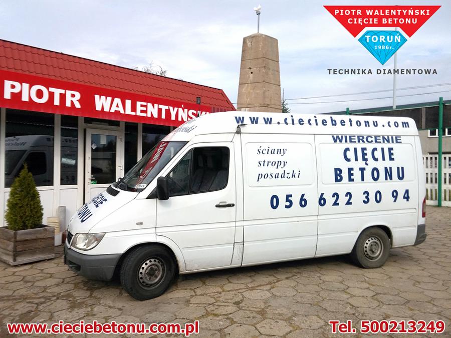 Biały samochód dostawczy z niebieskim napisem 'Cięcie Betonu' oraz numerem telefonu, zaparkowany na brukowanej powierzchni przed budynkiem z czerwonym dachem i szyldem 'Piotr Walentyński'.