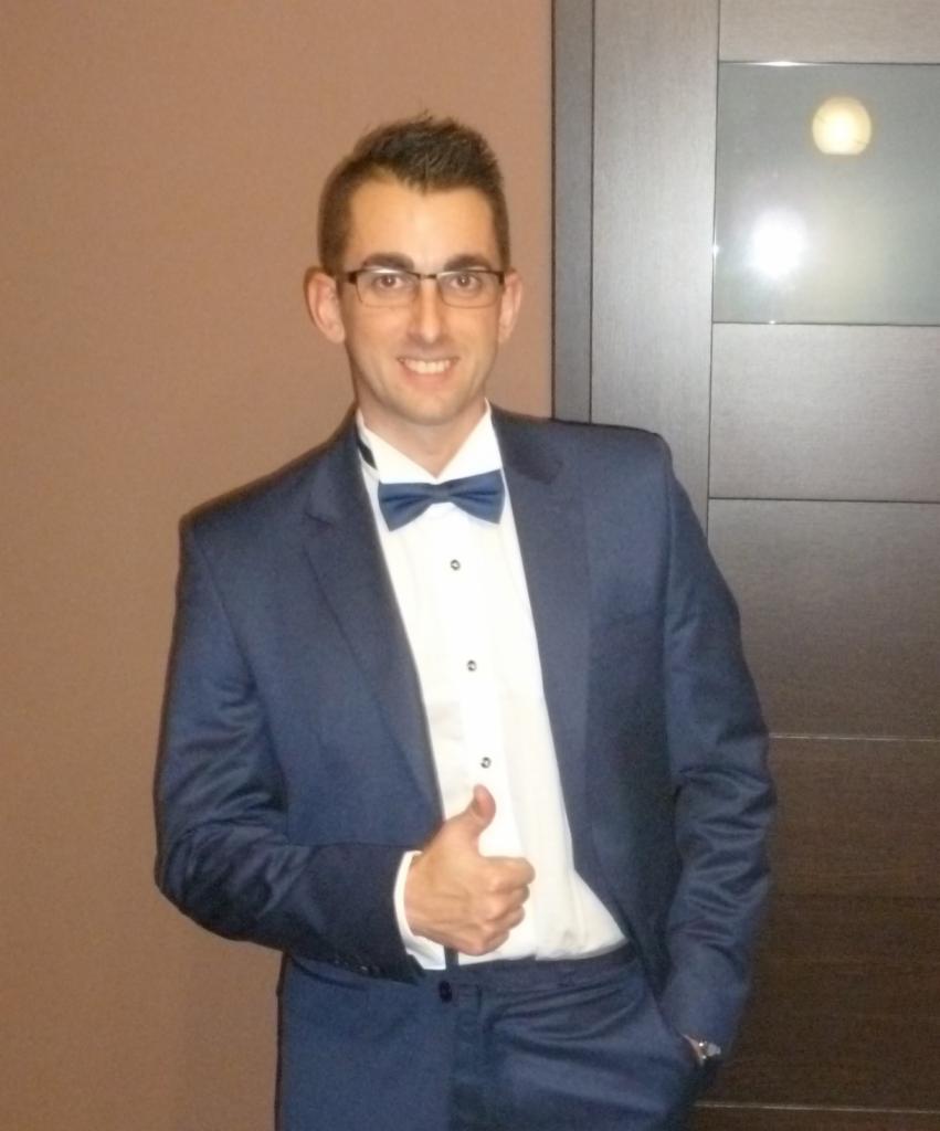 Uśmiechnięty mężczyzna w okularach, w granatowym garniturze z muchą, gestykuluje kciukiem w górę, stojąc na tle ściany i drzwi.