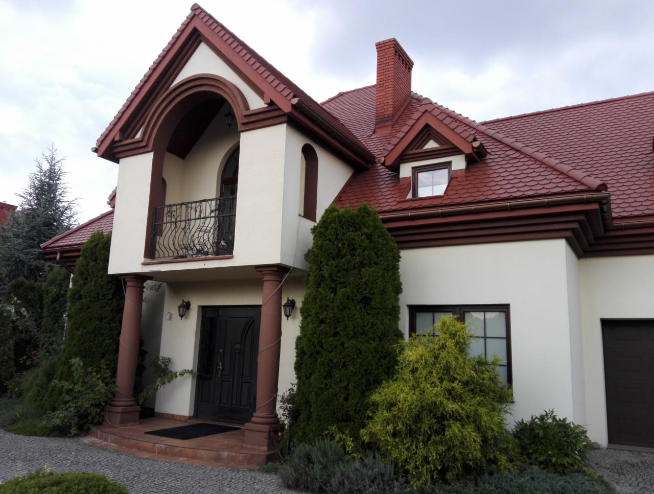 Elewacja domu jednorodzinnego z czerwonym dachem i elementami dekoracyjnymi w kolorze brązowym, widok z zewnątrz na front budynku z balkonem i kolumnami.