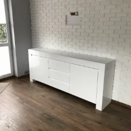 Biała, lakierowana komoda z trzema szufladami i dwojgiem drzwi, ustawiona przy ścianie z białej cegły dekoracyjnej. Widoczna podłoga z paneli imitujących ciemne drewno.