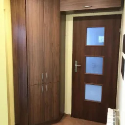 Wysoka, narożna szafa z drewnopodobną okleiną, dopasowana do wnęki obok drzwi z trzema kwadratowymi przeszkleniami, na tle żółtej ściany i brązowej podłogi.