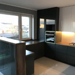 Nowoczesna kuchnia z zabudową w kolorze grafitowym i białym, z oknem wychodzącym na osiedle domów. Widoczne blaty z ciemnego drewna, piekarnik w zabudowie i płyta gazowa. Podłoga wyłożona jasnymi...