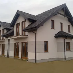 Nowoczesny, dwukondygnacyjny dom szeregowy z szarą dachówką i jasną elewacją z szarymi akcentami, widok z zewnątrz w pochmurny dzień.