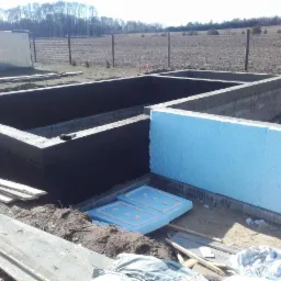 Surowe fundamenty domu z izolacją termiczną z niebieskiego styropianu, zabezpieczone czarną powłoką, widoczne deski szalunkowe i krajobraz pola w tle.