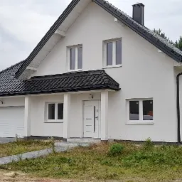 Nowoczesny, biały dom jednorodzinny z czarną dachówką, widok z zewnątrz, na trawniku widoczne pozostałości po budowie, chodnik z kostki prowadzi do wejścia.