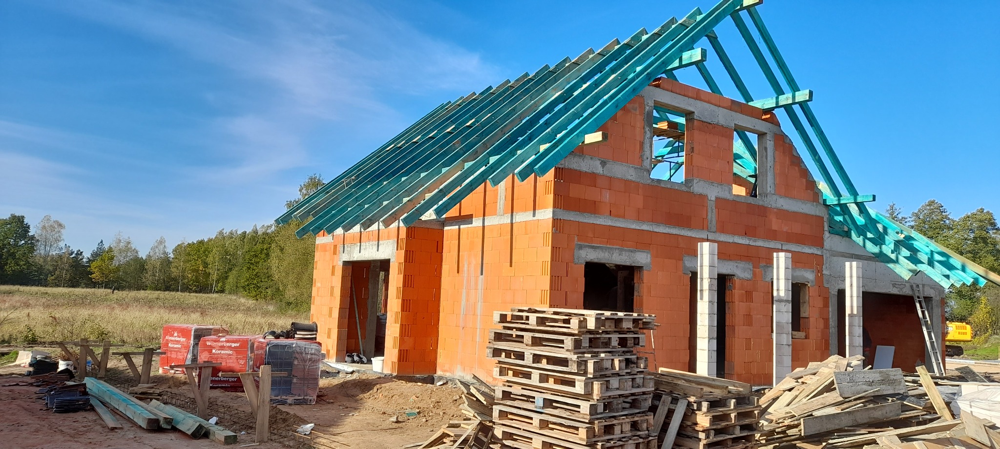 Surowy stan domu jednorodzinnego z czerwonej cegły, z widoczną konstrukcją dachu z impregnowanego drewna na tle błękitnego nieba, palety cegieł i drewniane palety na pierwszym planie.