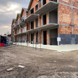 Surowy stan budynku wielorodzinnego w trakcie izolacji fundamentów; widoczna biała warstwa izolacyjna przy gruncie, szare płyty izolacyjne na ścianie i zaparkowany biały samochód dostawczy...