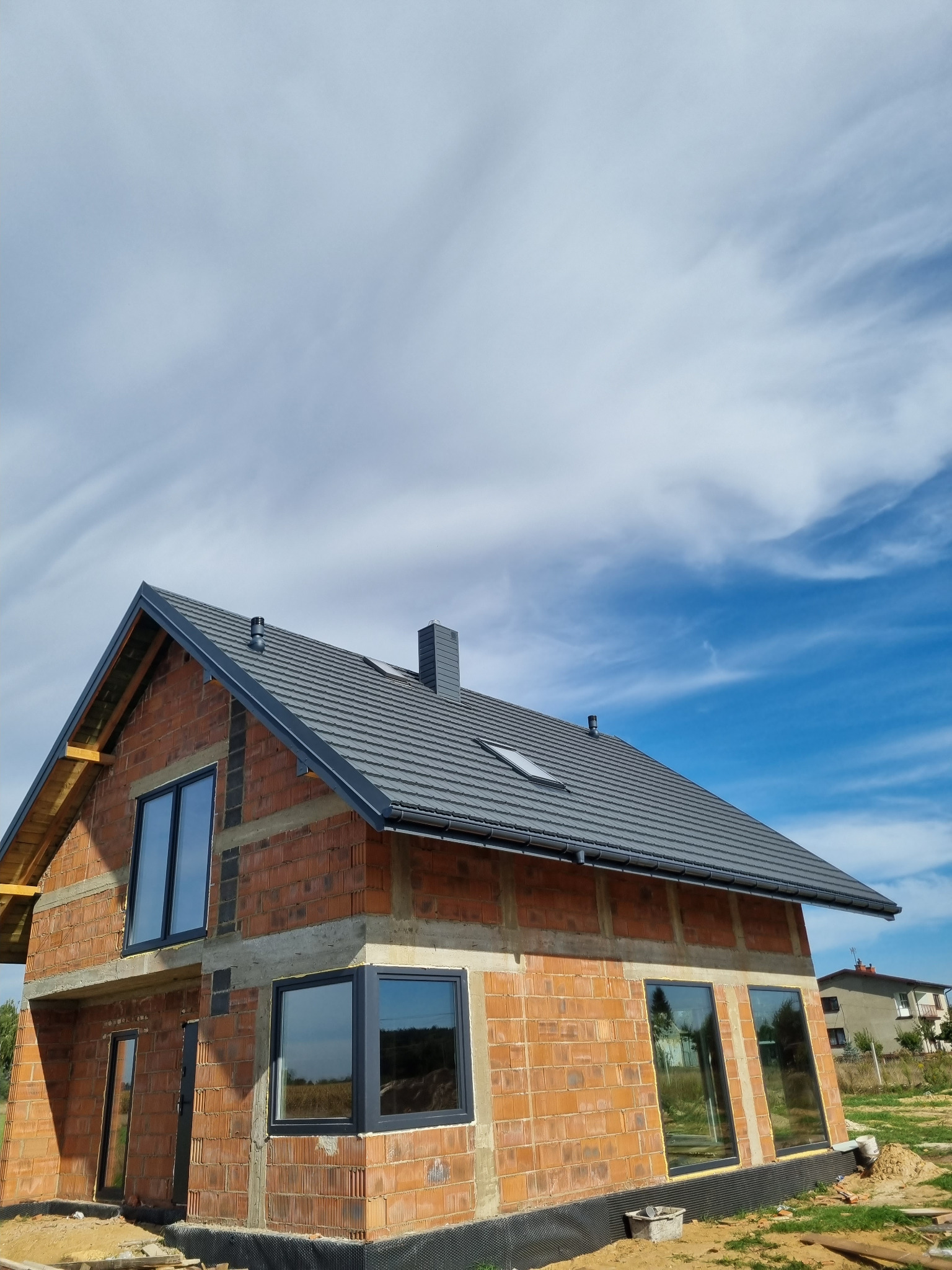Surowy dom z czerwonej cegły z szarym dachem i oknami, w trakcie budowy. Widoczne niebo z chmurami. Ujęcie z dołu, podkreślające bryłę budynku.