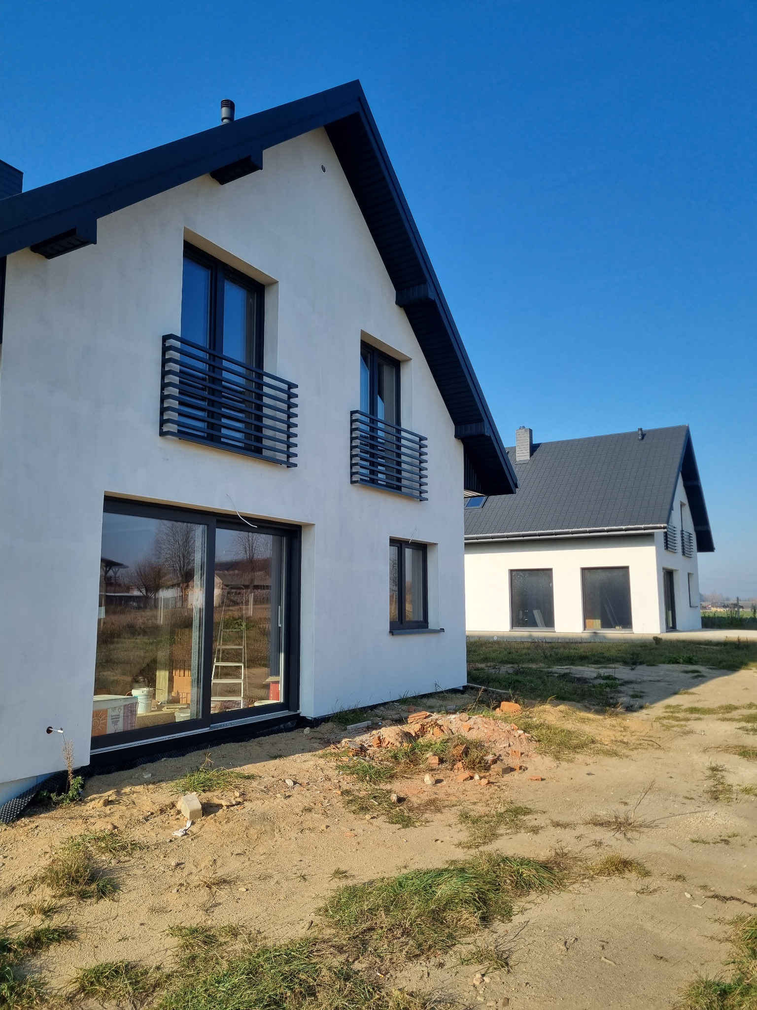 Dwa nowoczesne domy z białymi elewacjami i ciemnymi dachami w słoneczny dzień. Widoczne okna z czarnymi ramami i metalowe balustrady balkonowe. Teren wokół w trakcie zagospodarowania.