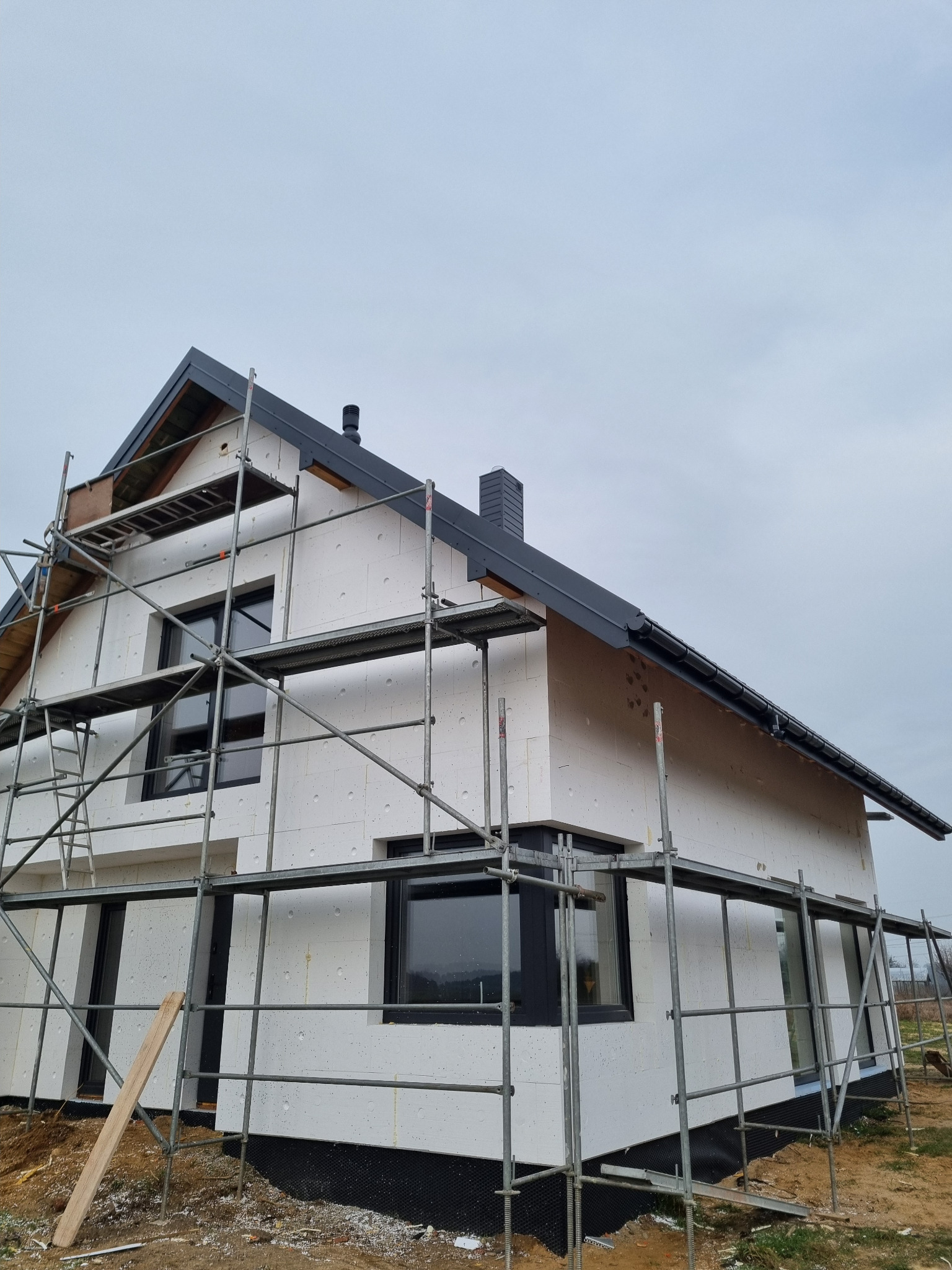 Ocieplony dom w trakcie budowy, z widocznymi rusztowaniami i czarną stolarką okienną na tle pochmurnego nieba. Detal: widoczna faktura styropianu.