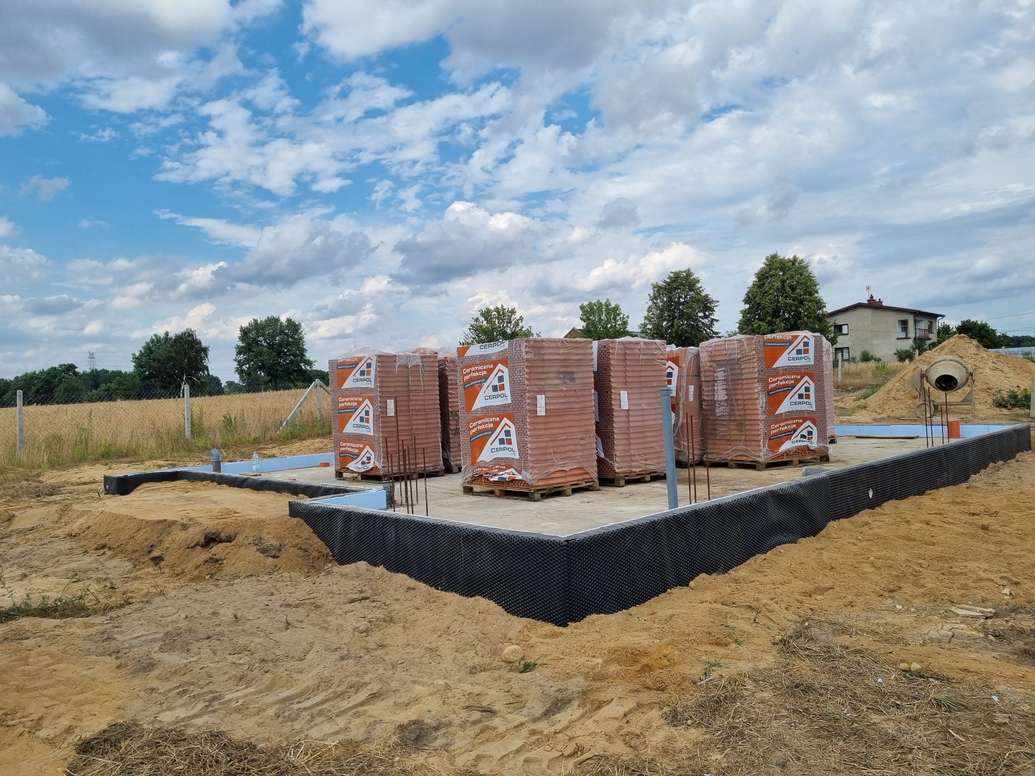 Fundament domu w trakcie budowy: zabezpieczona izolacja, styropian, wystające pręty zbrojeniowe, palety z cegłą CERPOL, betoniarka w tle, błękitne niebo.