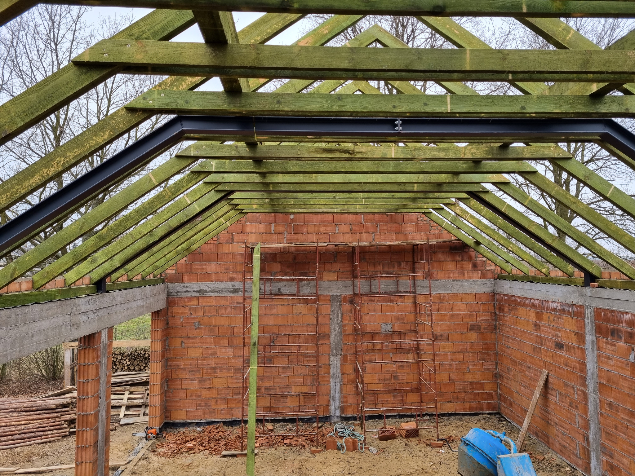 Wnętrze wznoszonego domu z czerwonej cegły z drewnianą konstrukcją dachu i stalowymi elementami. Widoczne rusztowania i narzędzia budowlane.