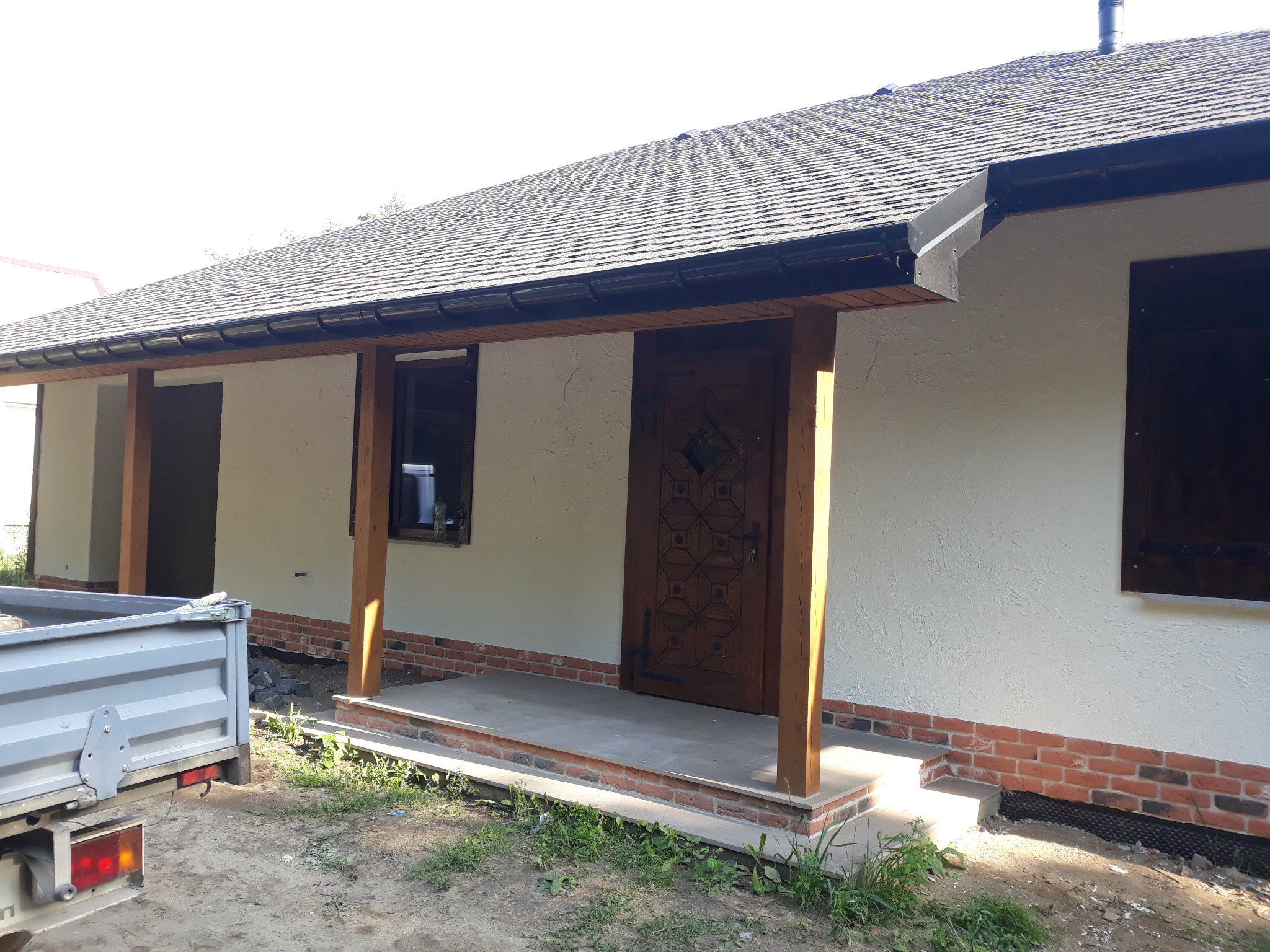 Wykończony dom z drewnianymi kolumnami werandy i nowym dachem z gontu bitumicznego. Widoczny fragment skrzyni ładunkowej samochodu dostawczego.