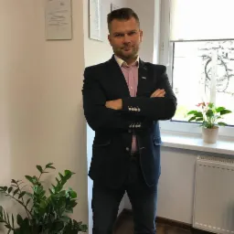 Mężczyzna w ciemnej marynarce i dżinsach stoi z założonymi rękami w biurze, za nim okno z kwiatami doniczkowymi, z lewej strony roślina doniczkowa.