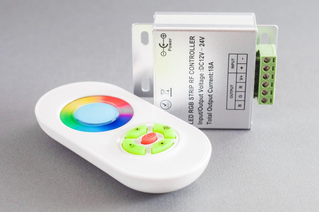 Srebrny sterownik LED RGB z zielonymi złączami i biały pilot z kolorowym pierścieniem dotykowym oraz zielonymi i czerwonymi przyciskami na szarym tle.