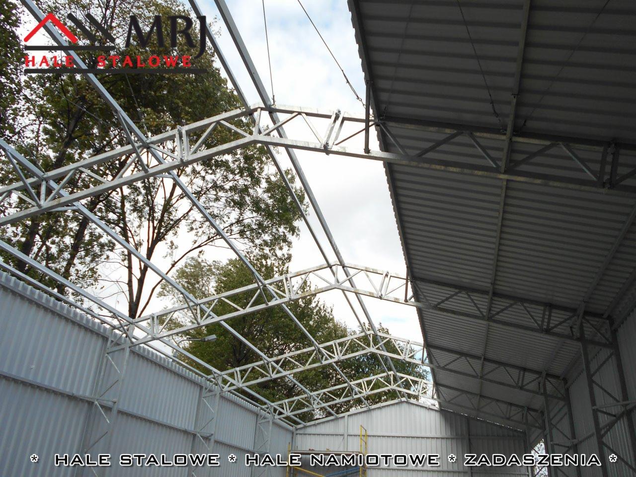 Wnętrze hali w trakcie budowy, widoczne elementy konstrukcji nośnej z profili stalowych, częściowo zamontowane poszycie dachu z blachy trapezowej, drzewa widoczne w tle przez niezabudowaną część...
