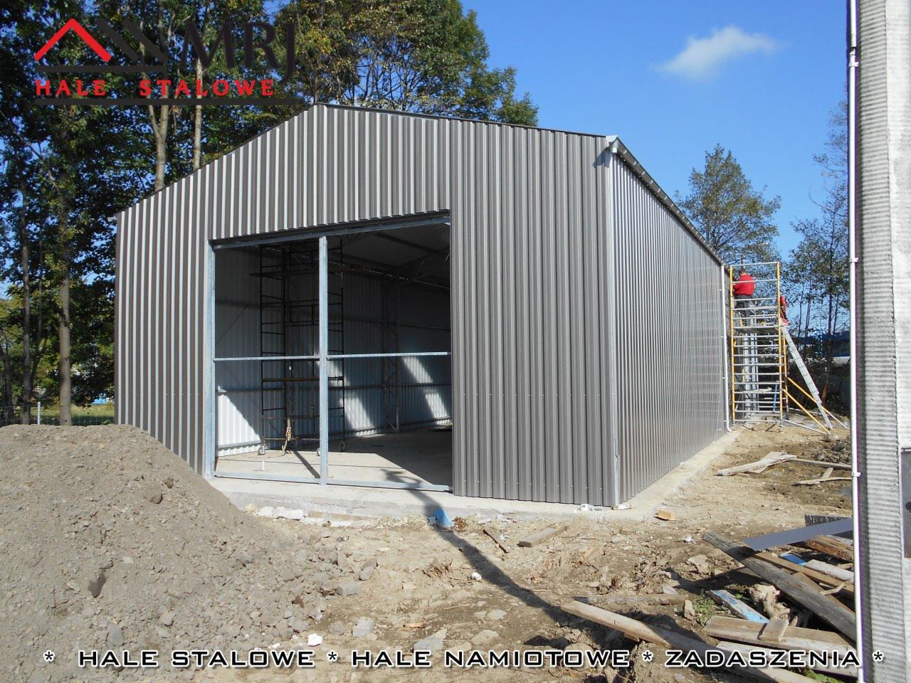Gotowa hala z blachy trapezowej T18 i T-35, z otwartym wjazdem, widoczne rusztowanie z pracownikiem na drabinie podczas montażu, na pierwszym planie pryzma ziemi.