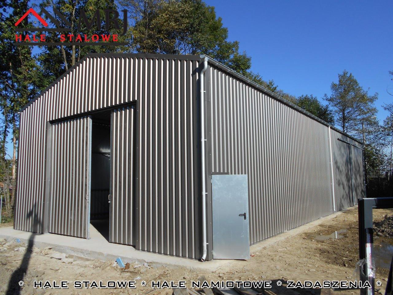 Gotowa hala stalowa z otwartymi przesuwnymi drzwiami i dodatkowym wejściem, pokryta blachą trapezową w kolorze srebrnym, na fundamencie betonowym, w tle drzewa i błękitne niebo.