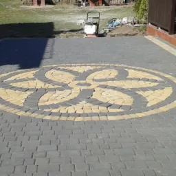 Ułożona kostka brukowa z dekoracyjnym wzorem kwiatowym w odcieniach szarości i beżu. W tle widoczny sprzęt budowlany i zieleń.