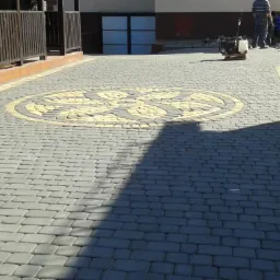 Ułożona kostka brukowa z dekoracyjnym wzorem w kształcie rozety. W tle widoczne budynki i maszyna do ubijania kostki.