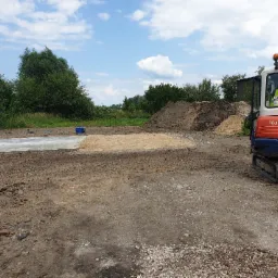 Przygotowany teren pod budowę szamba betonowego z widocznym wykopem, częściowo zasypanym piaskiem i betonową płytą fundamentową, obok stoi pomarańczowy mini-koparka Kubota, w tle drzewa i niebo...