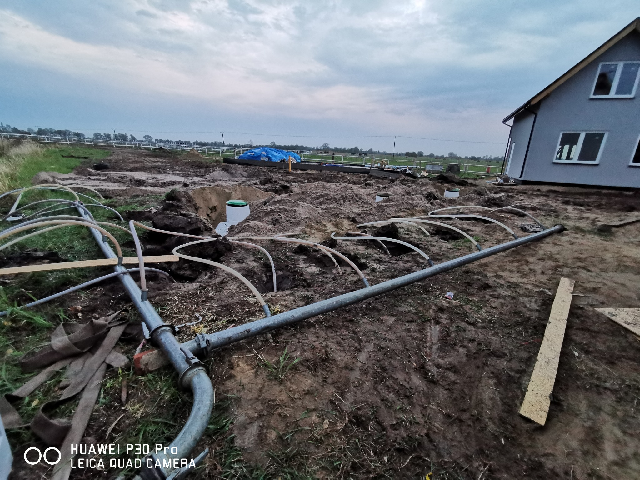 Instalacja drenażu rozsączającego na placu budowy z widocznymi rurami, ziemią i nowym domem w tle, pochmurne niebo.