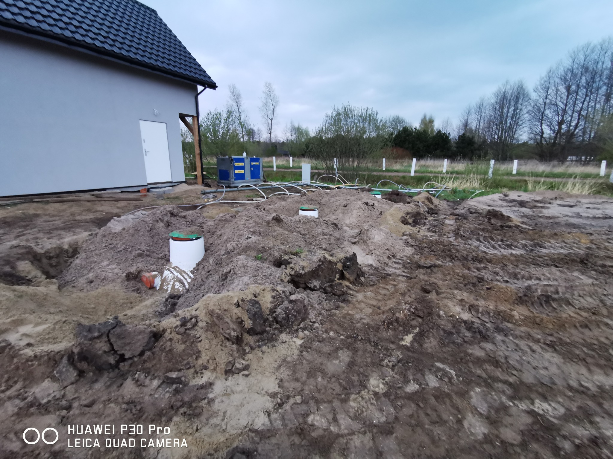 Teren budowy z wykopem pod instalację sanitarną, widoczne rury kanalizacyjne, studzienki rewizyjne, białe słupki ogrodzeniowe w tle oraz fragment budynku z szarą elewacją i białą stolarką.