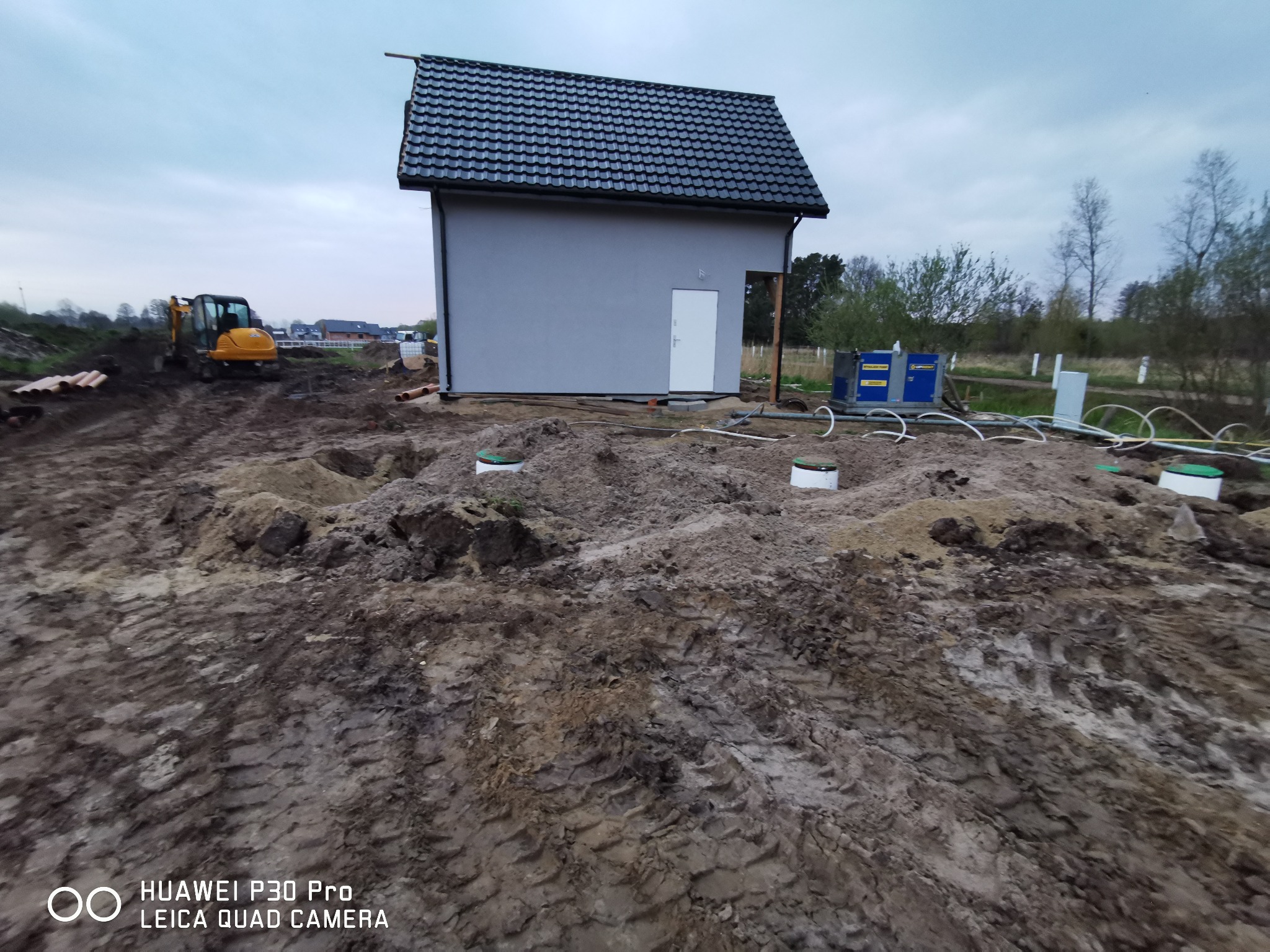 Budynek z szarą elewacją i ciemną dachówką, w tle mała koparka, widoczne białe rury i zielone pokrywy włazów na nierównym terenie z wykopami.