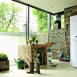 Nowoczesny, biały piec grzewczy Vaillant z niebieskim wyświetlaczem w jasnym pomieszczeniu z dużym oknem, obok ściana wyłożona kamieniem, wiklinowy stolik z dekoracjami i kalosze.