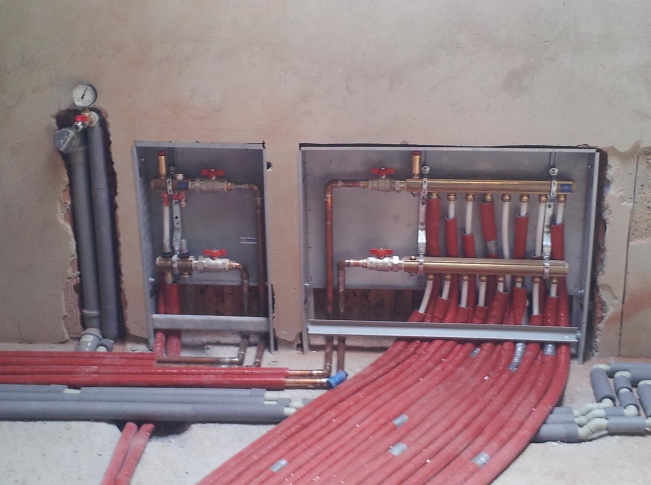 Rozdzielacze ogrzewania podłogowego z czerwonymi rurami w surowym wnętrzu, widoczne zawory i manometr na szarych rurach. Etap instalacji w trakcie prac wykończeniowych.