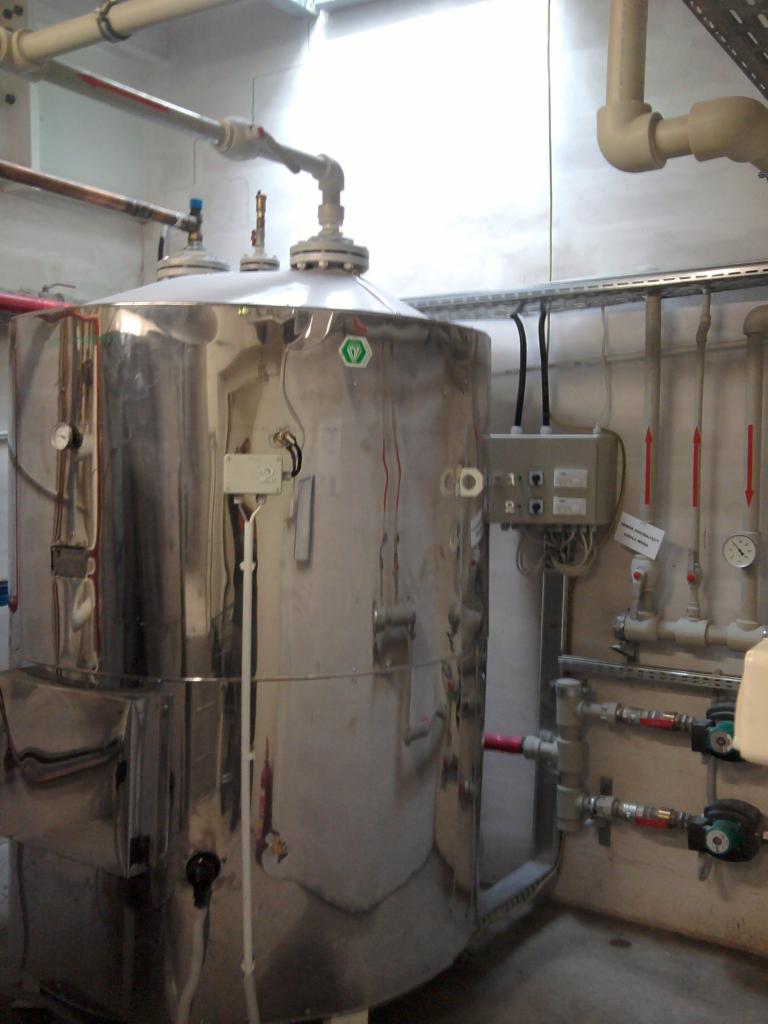 Bojler ze stali nierdzewnej z licznymi rurami i zaworami w kotłowni. Widoczne manometry, skrzynka elektryczna i instalacje w jasnym pomieszczeniu.