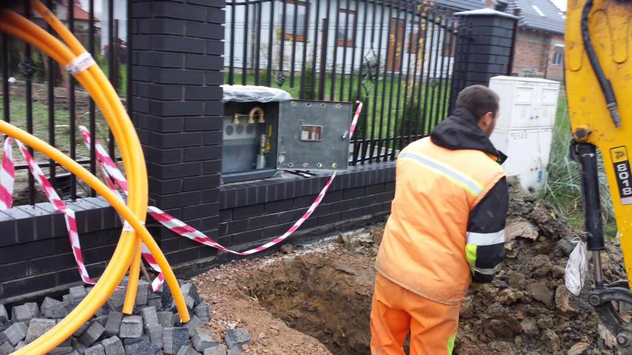 Instalacja gazowa w trakcie montażu: żółte rury gazowe przy wykopie i otwartej skrzynce z zaworami, pracownik w odblaskowej kamizelce i koparka JCB w tle.