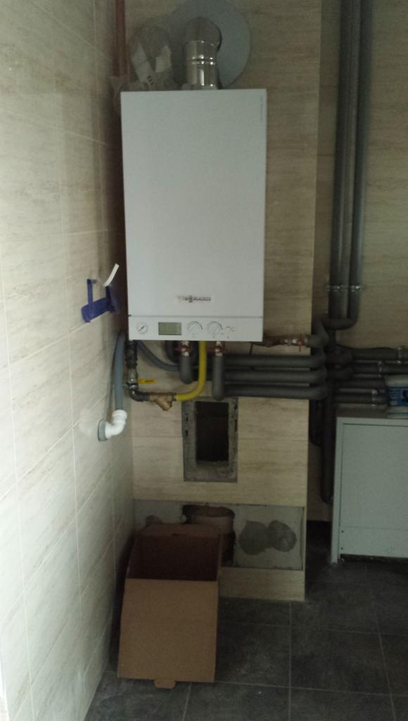 Biały piec gazowy Viessmann zamontowany na ścianie z beżowych płytek, widoczne rury i kartonowe pudło na podłodze. Ujęcie z bliska.