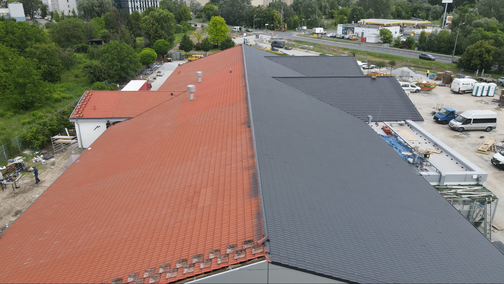Dach budynku podzielony na dwie części: jedna z czerwoną dachówką, druga z szarą dachówką. Widoczne kominy wentylacyjne. Na pierwszym planie fragment dachu z szarą dachówką, w tle budynki i zieleń.