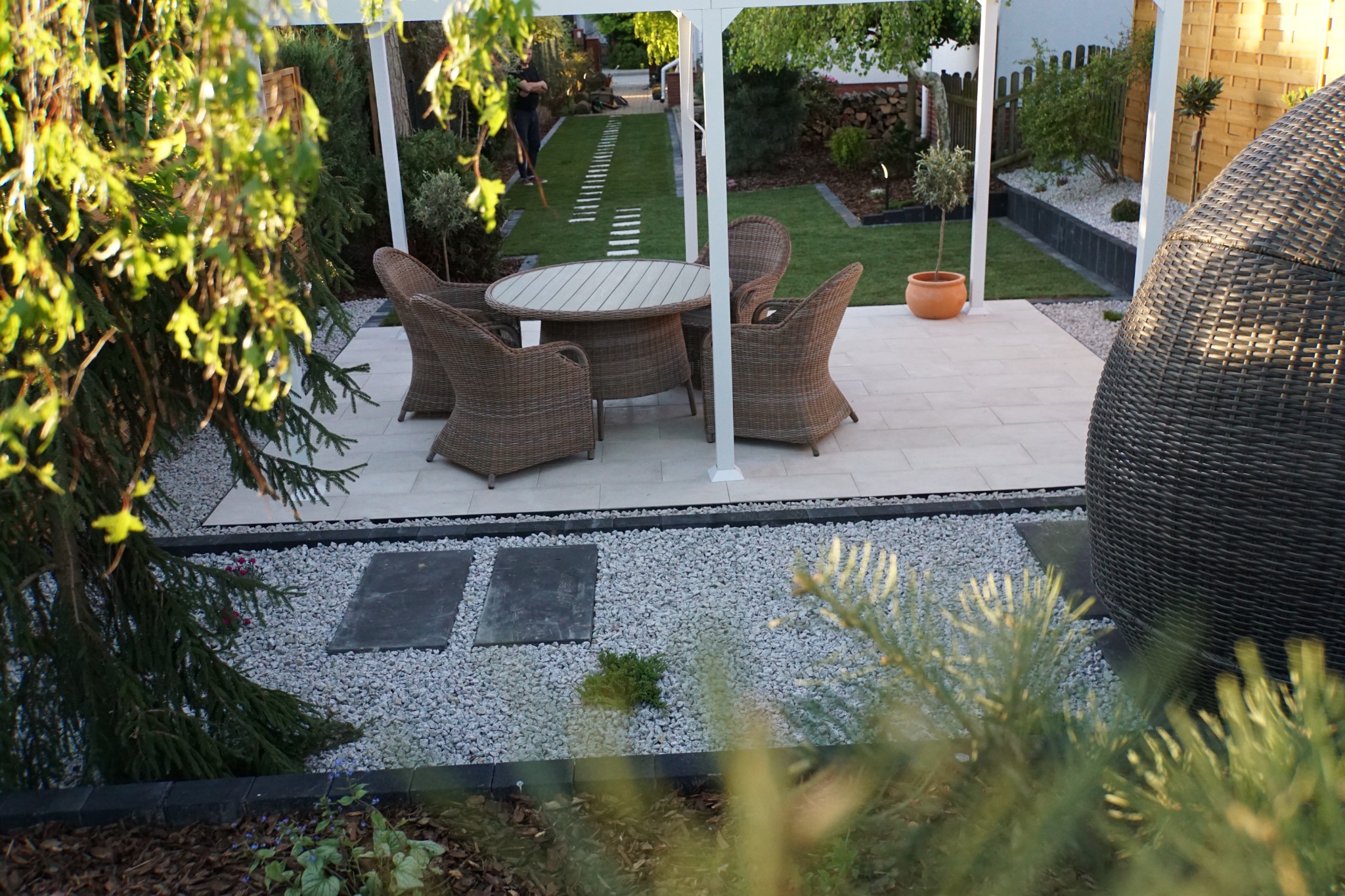 Zaciszny kącik wypoczynkowy w ogrodzie z meblami rattanowymi pod białą pergolą, ścieżka z płyt na trawniku w tle.