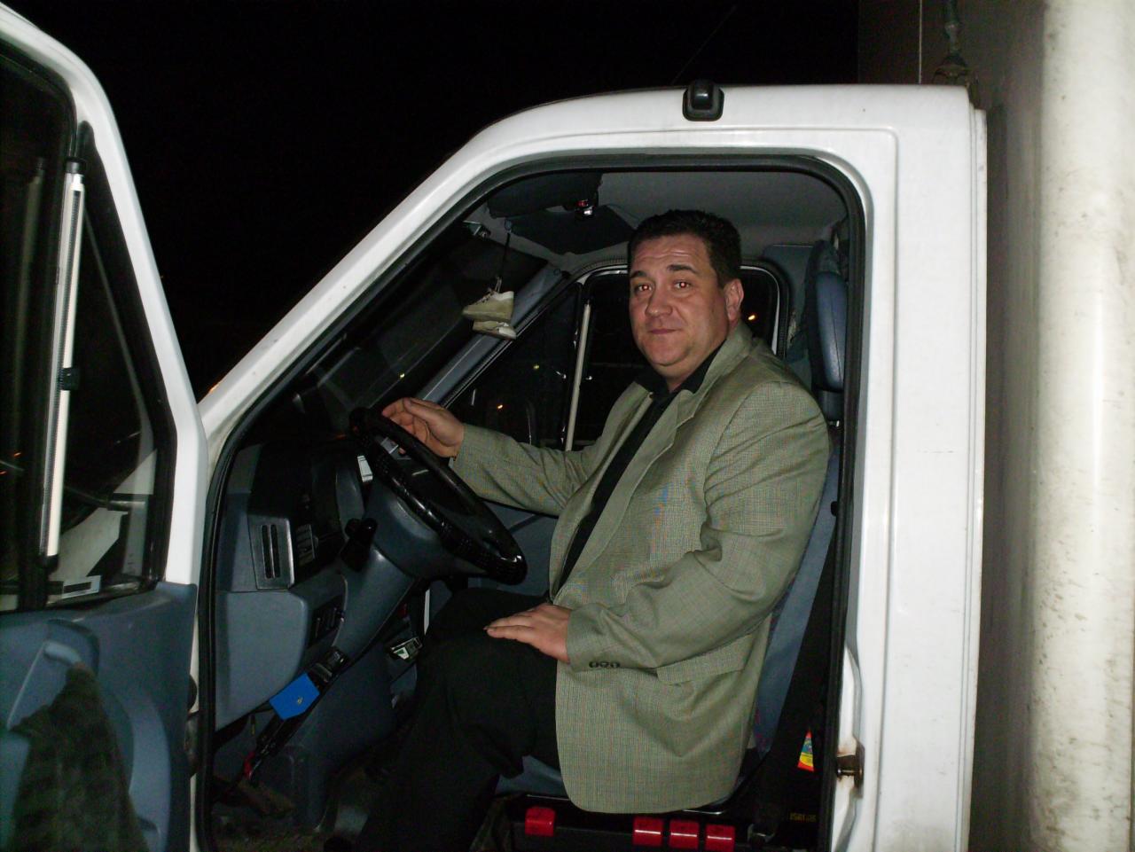 Kierowca w jasnym garniturze siedzi za kierownicą białego busa, gotowy do drogi. Widoczna deska rozdzielcza i otwarte drzwi pojazdu.