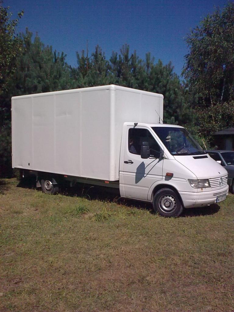 Biały bus dostawczy z zabudową kontenerową, zaparkowany na trawie w otoczeniu drzew. Widoczny fragment tablicy rejestracyjnej.