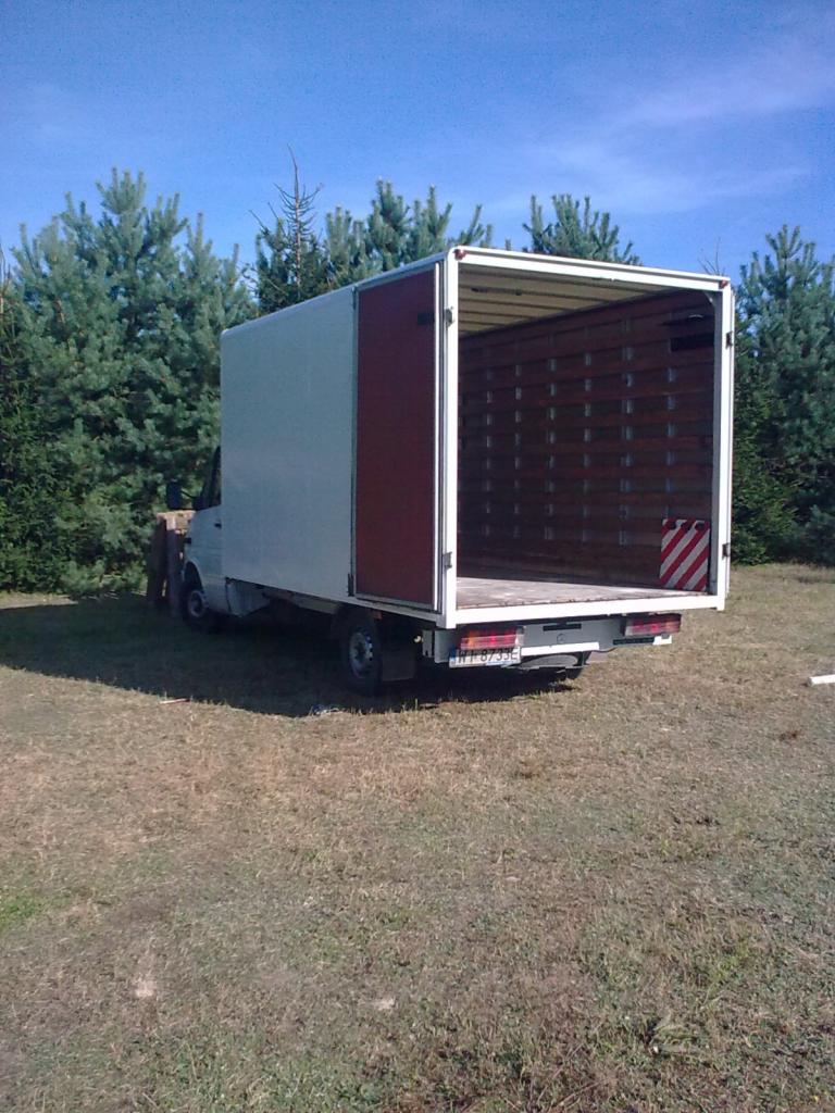 Pojazd dostawczy z otwartą przestrzenią ładunkową, zaparkowany na trawiastym terenie, widok z tyłu, błękitne niebo w tle.