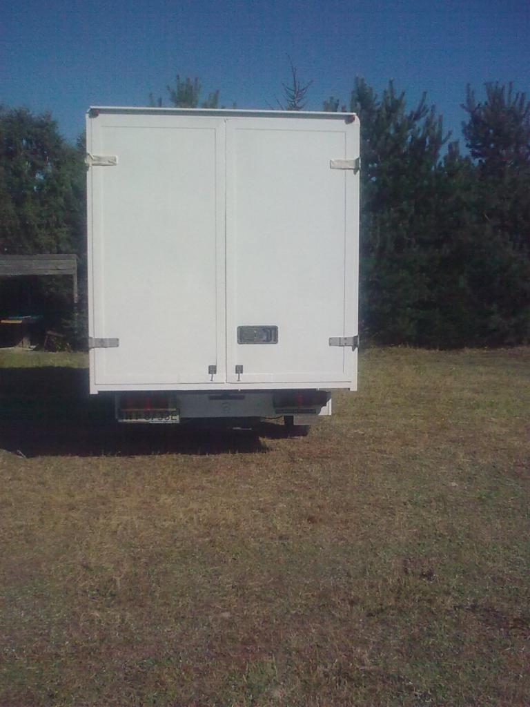 Tył białego busa z zamkniętymi drzwiami na tle trawy i drzew. Widoczny numer rejestracyjny. Ujęcie z bliska.