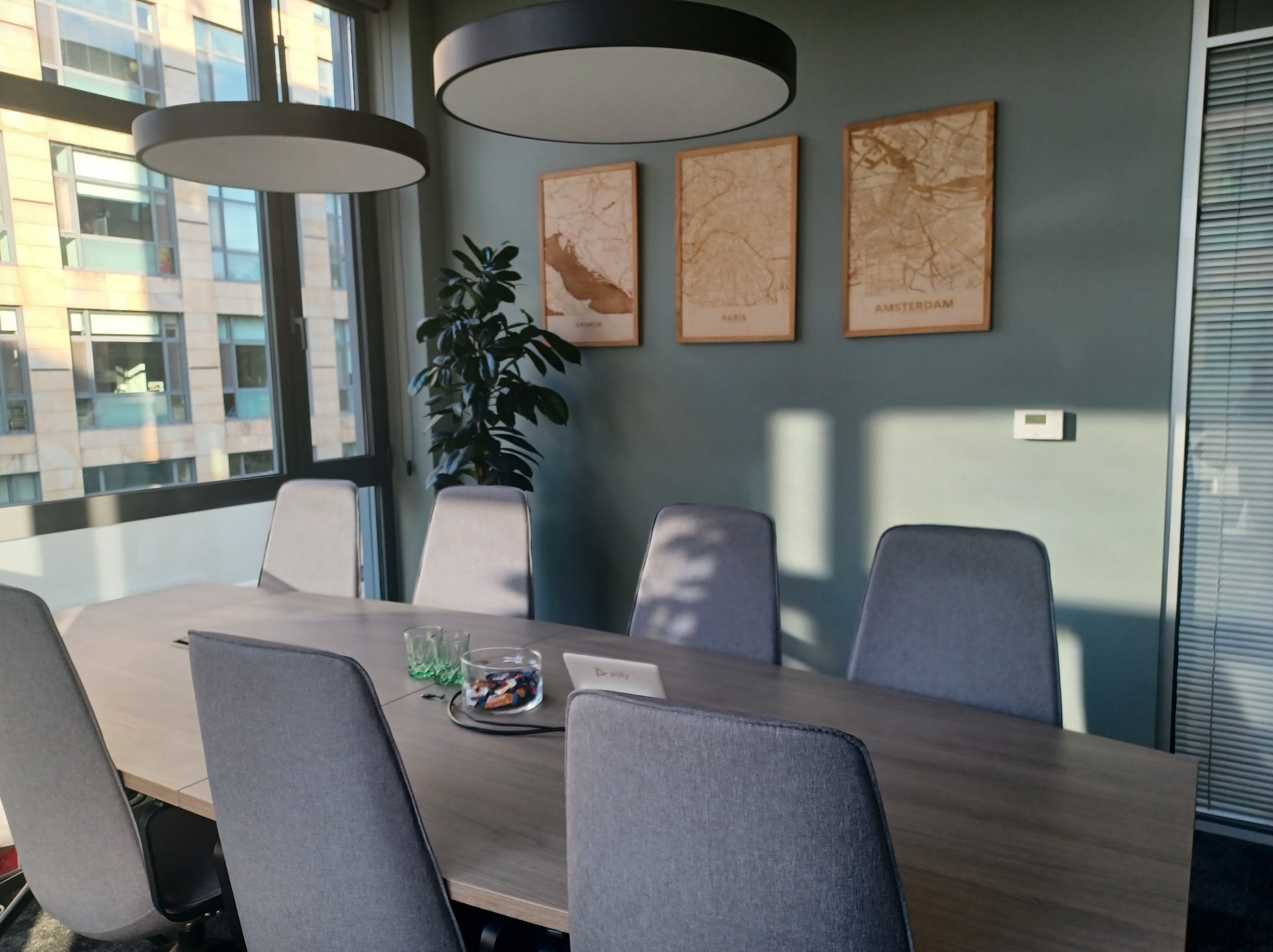 Nowoczesna sala konferencyjna z szarymi krzesłami, drewnianym stołem, minimalistycznymi lampami i mapami miast na ścianie. Widok przez okno na budynek.