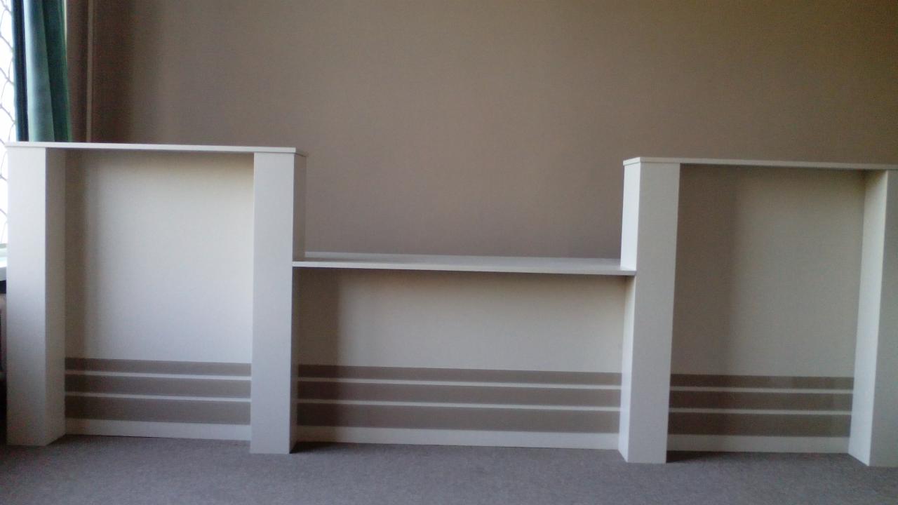 Biała zabudowa meblowa z półkami na beżowej ścianie, widok z przodu. Dolna część ściany zdobiona poziomymi pasami w odcieniach brązu.