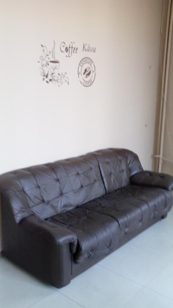 Ciemnobrązowa skórzana sofa z pikowaniem na tle ściany z naklejkami 'Coffee Káva' i symbolem espresso. Widok z perspektywy.