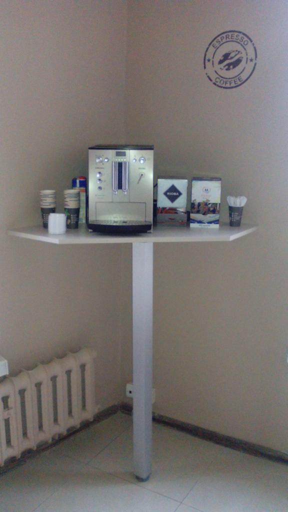 Stylowy kącik kawowy z ekspresem, kubkami i zapasem kawy na białym stoliku. Na ścianie logo espresso. Widoczny grzejnik. Ujęcie z perspektywy podłogi.