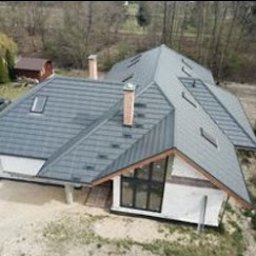 WESKAD-BUD Budownictwo - Nowoczesny dom jednorodzinny w trakcie budowy, widoczna konstrukcja dachu z szarej blachy trapezowej, fragment elewacji z oknami oraz komin z cegły. W tle drzewa i mały budynek gospodarczy.