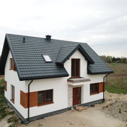WESKAD-BUD Budownictwo - Nowo wybudowany, dwukondygnacyjny dom z ciemnoszarą dachówką, białymi ścianami i drewnianymi akcentami, otoczony ziemią i zielenią, w pochmurny dzień.