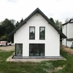 Nowoczesny, biały dom jednorodzinny z ciemnymi oknami i dachem dwuspadowym na tle drzew. Minimalistyczny design, świeżo wykończona elewacja, widoczny fragment samochodu dostawczego.