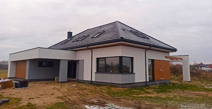 Nowoczesny dom jednorodzinny z płaskim dachem, wykończony w bieli i szarości z elementami drewnopodobnymi, widoczne okna dachowe i teren wokół w trakcie zagospodarowywania.
