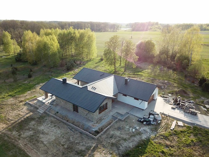 Widok z drona na parterowy dom z szarej cegły z ciemną dachówką, w trakcie wykończenia terenu, z widocznymi fundamentami i stertą materiałów budowlanych na podjeździe, na tle zielonego pola i lasu.